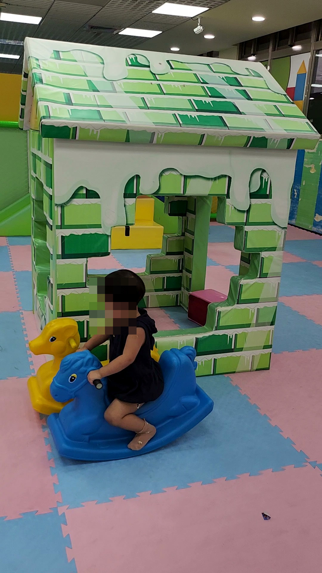 孩子坐在搖搖馬上,後面是綠色磚牆造型小屋
